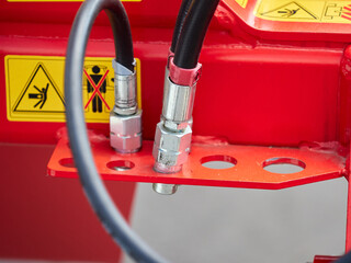Close-up of two hydraulic hose fittings attached to a red industrial machine, with visible safety warning labels in the background.
