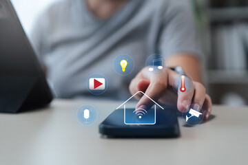 Hand using smartphone to control smart home devices; icons for light, music, thermostat, camera and...