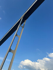 A tall bridge looms with its steel framework rising high against a vibrant blue sky. Fluffy white...