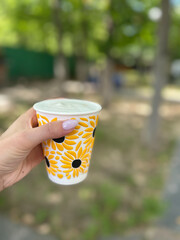 A hand grasps a colorful cup adorned with flowers, surrounded by greenery on a bright day. The...