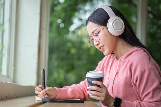 A beautiful Asian woman wearing glasses and headphones is sitting in a cafe thinking and taking notes.