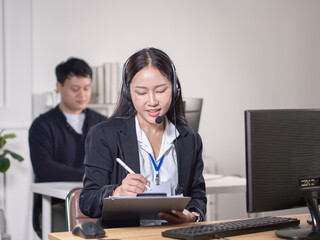 An Asian woman in a suit and headset is taking notes at a computer. Look like a call center
