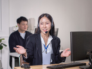 An Asian woman in a suit and a headset is explaining in front of a computer, similar to a call...