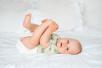 a baby boy plays with his legs while lying on his back on a white bed at home, a smiling little...