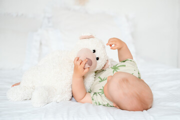 a baby boy plays with a soft teddy bear lying on his back on a white bed at home