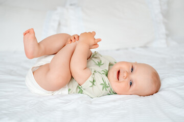 a baby boy plays with his legs while lying on his back on a white bed at home, a smiling little...