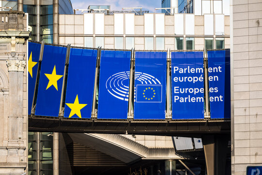Wide view of the European Parliament's Altiero Spinelli building entrance in Brussels, Belgium, framed by a large curved pedestrian bridge adorned with EU-themed banners.