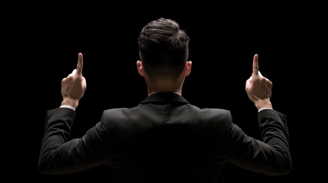 Leadership Pose: A man in a dark suit stands with his back to the camera, fingers pointed upwards, symbolizing guidance, direction, and authority in a dramatic spotlight against a black background.