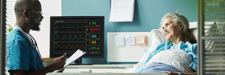 Header of senior Caucasian woman lying in hospital bed connected to oxygen tube, Black male doctor...