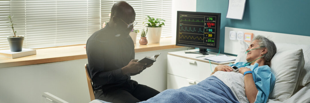 Senior Caucasian woman lying in hospice bed receiving oxygen therapy, Black male chaplain sitting beside bed reading from Bible, medical monitor displaying vital signs in background