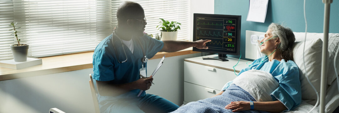 Header of Black male doctor explaining heart monitor results to senior Caucasian female patient lying in hospital bed with nasal cannula, medical equipment in background, woman listening attentively - Powered by Adobe