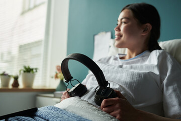 Caucasian young pregnant woman reclining in bed in maternity hospital holding wireless headphones,...