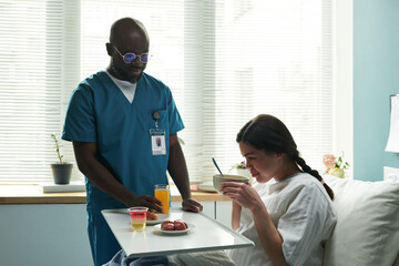 Black middle aged male nurse serving food and drinks to young pregnant Caucasian woman sitting in...