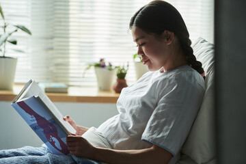 Caucasian young pregnant woman sitting on bed reading maternity book, natural light coming through...