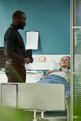 Middle aged Black male priest with Bible standing beside hospital bed talking to senior Caucasian...