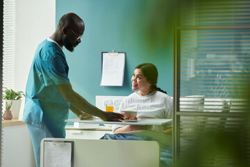 Middle aged Black male nurse wearing medical scrubs serving food tray to young pregnant Caucasian...