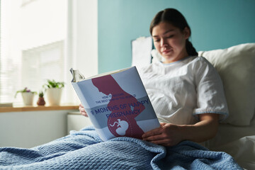 Caucasian young adult woman sitting in hospital bed reading pregnancy book, holding book with both...