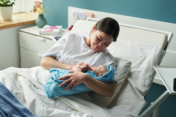 Caucasian young woman lying in hospital bed holding newborn baby, smiling and looking at infant,...