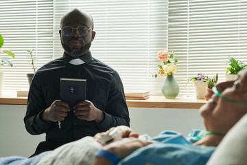 Black mature male chaplain wearing clerical collar, holding bible standing beside hospital bed,...