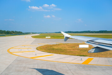 View from an aircraft window shows the wing and an airport runway on a sunny day. Grass and trees...