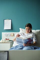 Young pregnant Caucasian woman sitting in hospital bed reading maternity manual book, wearing...