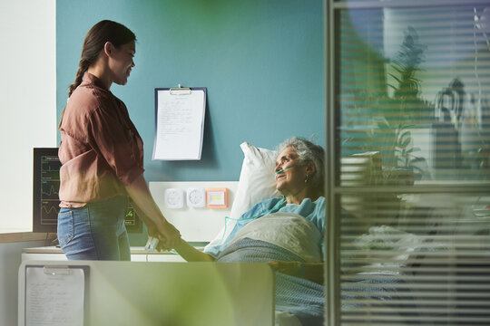 Young Caucasian woman standing beside hospital bed holding hand of senior female family member lying with oxygen tube, both smiling and making eye contact in medical setting