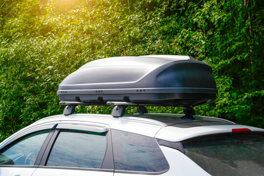 A white vehicle has a roof box securely mounted on top. The container appears to be made of durable plastic, and is being used for travel and additional storage. Trees are visible in the background