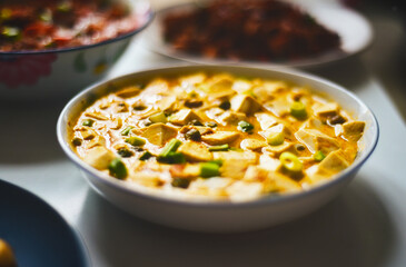 A bowl of creamy, yellowish soup with tofu and green vegetables, served alongside other dishes.