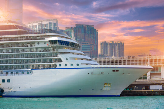 A majestic white cruise ship is docked at a pier in Hong Kong. The city skyline and vibrant sunset create a breathtaking backdrop