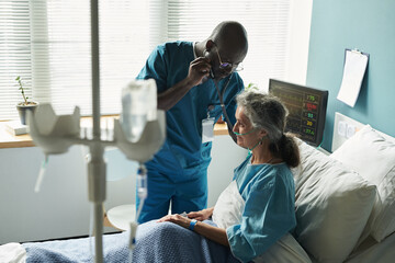 Middle aged Black man doctor examining senior Caucasian woman patient sitting in hospital bed using...