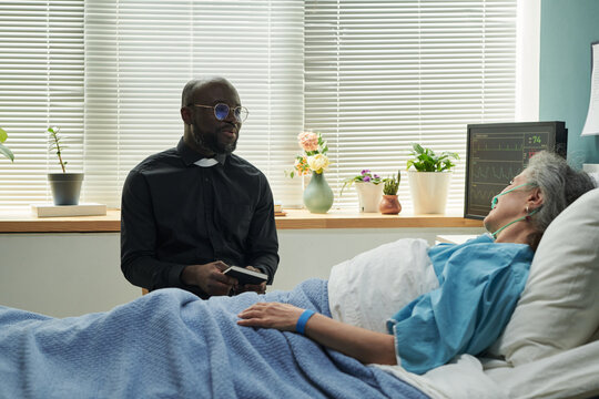 Middle aged Black male chaplain wearing clerical collar sitting beside senior Caucasian woman lying in hospital bed with oxygen tube, holding book and talking, medical monitor in background