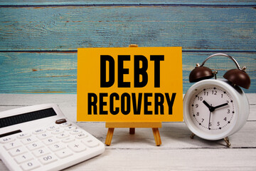 Debt Recovery text written on paper card with calculator and alarm clock on wooden background