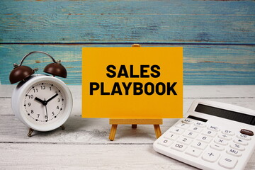 Sales Playbook text written on paper card with calculator and alarm clock on wooden background