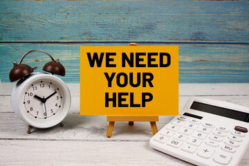 We Need Your Help text written on paper card with calculator and alarm clock on wooden background