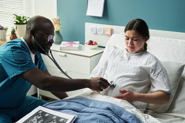 Black male doctor examining pregnant Caucasian young woman with stethoscope on baby belly in...