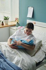 Young Caucasian woman sitting in hospital bed in maternity ward, holding newborn baby in arms,...