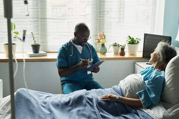 Black middle aged male doctor sitting beside hospital bed, writing on clipboard, while senior Caucasian woman patient lying under blanket, connected to medical monitor in patient room