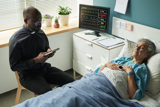 Black middle aged male chaplain sitting beside hospital bed using smartphone while senior Caucasian woman lying unconscious with oxygen tube, providing spiritual support and praying