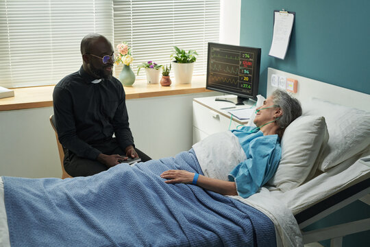 Middle aged Black male priest sitting beside hospital bed talking to senior Caucasian woman with oxygen tube lying under blanket, medical monitor displaying vital signs in background