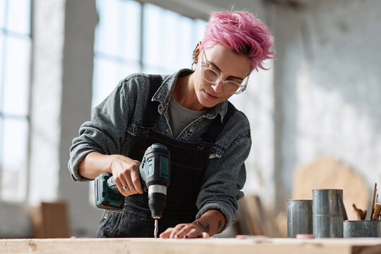 Focused artisan with pink hair uses a drill on wood in a bright workshop. Symbol of craftsmanship, DIY, and female empowerment. Ideal for construction or renovation projects.