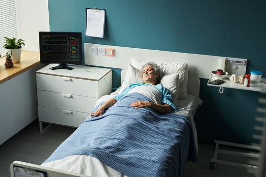 Senior Caucasian woman lying in hospital bed with medical monitor displaying vital signs, resting with eyes closed, wearing patient gown, medical equipment visible in background