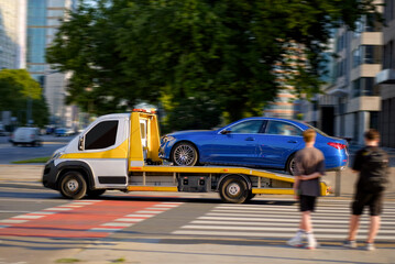 Yellow Cabin Tow Truck Transporting Damaged Luxury Premium C-Class Blue Car Through City Street In...