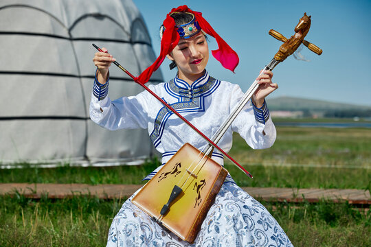 A Mongolian female musician in traditional Mongolian attire playing the morin khuur (horsehead fiddle) in front of a yurt.