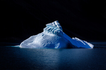 Blue iceberg floats on the deep waters of Dickson fjord, Northeast Greenland National Park. Dark background with reflection showing in the gentle ripples of the cold blue waters.