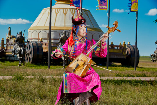 A Mongolian female musician in traditional Mongolian attire playing the morin khuur (horsehead fiddle) in front of a yurt.