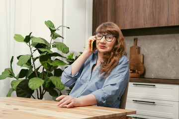 Beautiful elderly woman talking emotionally on smartphone in yellow case in stylish modern kitchen