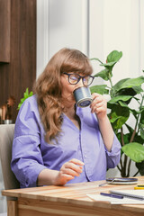 Old woman drinking coffee while busy studying with books and notebooks in stylish modern apartment