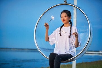 Fototapeta premium A cute girl setting off sparklers on a transparent swing by the lake at dusk.