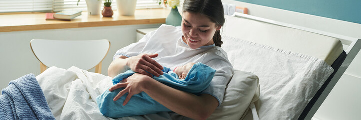 Header of Caucasian young woman sitting in hospital bed cradling newborn baby in arms, smiling and...