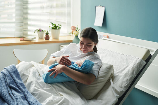 Joyous young Caucasian woman lying in hospital bed holding newborn baby, smiling and admiring infant with love, hospital room background with window and plants visible